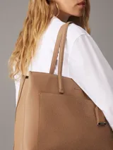Cartera tote Calvin Klein color tostado, de tamaño mediano, confeccionada en cuero sintético granulado con textura de logo monograma en el bolsillo delantero. Tiene asas fijas, cierre magnético, bolsillo exterior, compartimento interior con cremallera y colgante extraíble con logo.