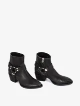 Bota corta de cuero negro con punta estilizada y taco bajo. Presenta un detalle de arnés desmontable alrededor del tobillo con argolla metálica y tachas plateadas.