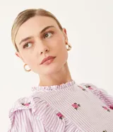 Blusa de algodón a rayas verticales blancas y lilas, con bordado de rosas rosadas. Tiene cuello redondo con volados, mangas largas y volados en los hombros.