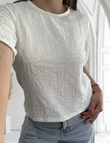 Blusa blanca de manga corta con cuello redondo y textura arrugada o plisada con patrón de pequeños cuadros.