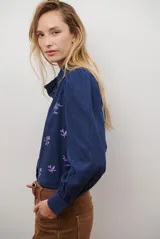 Camisa azul marino con estampado floral en color lila, cuello mao con volantes, cierre frontal de botones y mangas largas abullonadas con puños elásticos.