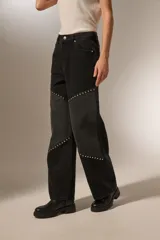 Pantalón de jean negro lavado, tiro alto, corte recto y ancho. Presenta paneles diagonales en la parte delantera de las piernas adornados con tachas metálicas plateadas.