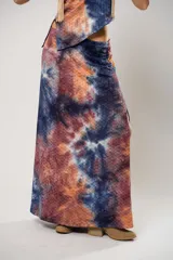 Conjunto de falda larga y top asimétrico con estampado tie-dye en tonos azul, naranja y marrón. El top es de un solo hombro y tiene un collarín con flecos dorados.