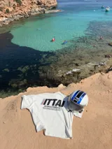 Remera color crema de corte recto y mangas cortas, con estampado en la espalda con el logo "ITTAI" en negro con líneas rojas, azules y blancas, y la leyenda "AN IBIZA PRODUCE. Ittai Stars Department Gallery - Ibiza Edition. If you're in Ibiza, 112 is not an option.".