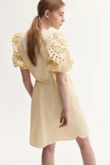 Vestido color crema a la rodilla, de tejido liviano de algodón con cuello redondo, botones ocultos adelante y cinturón fino extraíble. Mangas al codo abullonadas en broderie con puños elásticos.