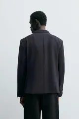 Blazer negro de corte relaxed fit confeccionada en tejido con lana. Cuello con solapas de muesca y manga larga acabada en puño con botones. Bolsillos de vivo en pecho y de solapa en cadera. Detalle de bolsillo interior. Bajo con aberturas en espalda. Cierre frontal de botonadura.