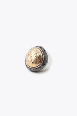 Maxi anillo metálico plateado con forma ovalada y pieza incrustada labrada dorada a contraste. Detalle de aplicación de brillos negros alrededor de la pieza dorada.