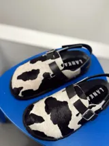 Zuecos con estampado de vaca en blanco y negro, con hebilla ajustable y suela plana.
