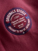 Buzo de manga larga color rojo tibetano, con cuello alto y cierre frontal. Presenta un fit suelto, interior afelpado y una estampa circular en el pecho con el texto "Originals Studio 1990".