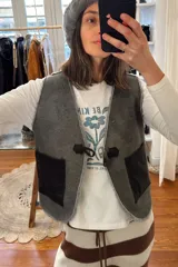 Chaleco marrón de cuero con interior de corderito color crema, cierre con gancho metálico y bolsillos frontales de corderito.