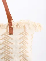 Bolso tipo tote color beige con diseño geométrico en blanco y flecos en la parte superior. Tiene doble asa de hombro marrón.