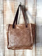 Bolso de mano o de hombro color marrón cobrizo con acabado metalizado y textura brillante. Posee dos asas cortas de cuero marrón oscuro y un bolsillo exterior frontal.