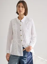 Camisa blanca de lino con cuello clásico, cierre frontal con botones marrones y mangas largas con puños abotonados. Presenta detalles de frunces verticales en el frente.
