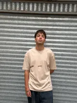 Remera oversize color beige con cuello redondo y manga corta. Presenta un pequeño corte o rasgadura en la parte delantera izquierda y estampados minimalistas en color marrón oscuro: la palabra "OSTEN" y un símbolo gráfico.