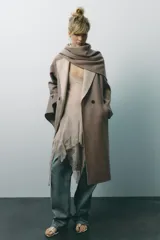 Abrigo largo color beige confeccionado en lana. Presenta cuello solapa con bufanda desmontable, manga larga, bolsillos laterales y cierre frontal cruzado con botones.