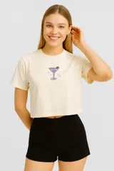 Remera cropped color crema con estampado de una copa de martini violeta y estrellas rosadas, de corte holgado y cuello redondo.