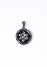 Dije redondo plateado de acero quirúrgico con diseño de flor de seis pétalos sobre fondo negro.