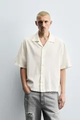 Camisa de denim de algodón, color beige claro, con corte relajado, cuello bowling y manga corta. Presenta cierre frontal con botones a presión y un pequeño bordado a tono en el pecho.