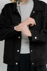 Chaqueta color beige de corte recto, estilo camisa, confeccionada en gamuza con elastano. Presenta cuello camisero, cierre frontal con botones metálicos, bolsillos grandes en el frente y charreteras en los hombros.