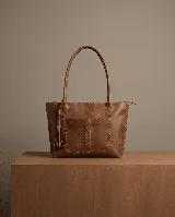 Cartera tote de cuero color marrón claro, con doble asa de hombro, bolsillo frontal con pespuntes decorativos y cierre superior. Incluye forrería y un dije de borla de cuero en una de las asas.