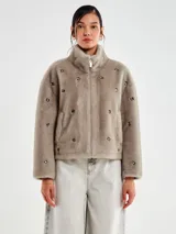 Chaqueta corta de pelo sintético color beige topo, con cuello alto y cierre frontal de cremallera plateada. La prenda está decorada con apliques metálicos circulares incrustados por toda la superficie.