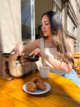 Cartera tipo tote de cuero color beige, con doble asa corta marrón y cierre superior con cremallera. Tiene un bolsillo frontal con cremallera y forro interior de tela con un bolsillo.