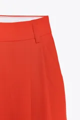 Pantalón de vestir color naranja, de tiro alto y pernera ancha. Presenta doble pliegue frontal, trabillas en la cintura, bolsillos laterales y falsos bolsillos de vivo en la espalda.