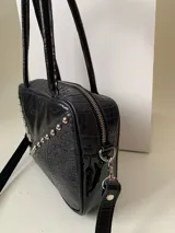 Cartera rígida negra de cuero con tachas plateadas y tres combinaciones de cuero diferentes que aportan textura. Tiene herrajes y cierre metálico plateado marca YKK. Cuenta con un asa fina para llevar al hombro o en la mano y una correa larga extra regulable.