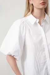 Camisa blanca de algodón con cuello camisero y mangas cortas abullonadas. Presenta un corte relajado y cierre frontal de botones.