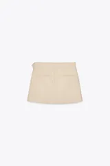 Falda pantalón color beige de tiro medio con diseño cruzado y cierre con botones. Cuenta con bolsillos falsos en la parte delantera y trasera, y cierre lateral con cremallera oculta.