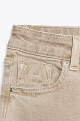 Pantalón de jean color beige, de tiro alto, corte ajustado en la cadera y muslos, y acampanado desde la rodilla. Tiene cinco bolsillos y cierre frontal con cremallera y botón.