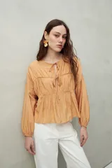 Blusa color beige de caída fluida con cuello redondo y lazos frontales. Diseño con cortes horizontales y frunces, mangas largas con puños elastizados.
