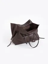 Bolso tote negro de cuero vacuno con textura granulada. Presenta cierres funcionales en los laterales para expandir el espacio y dos asas de mano. Incluye un tag colgante con aplique metálico y un tirador de cremallera con forma de figura.