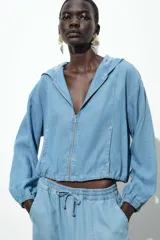 Chaqueta celeste de denim con capucha, cierre frontal con cremallera, bolsillos delanteros y mangas largas con puños elásticos.