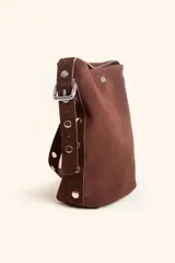 Cartera tipo balde de gamuza color marrón, con correa ajustable decorada con tachas metálicas plateadas y logo grabado en la parte frontal.