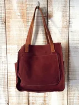 Bolso de mano o bandolera color marrón rojizo, con asas cortas de cuero marrón y correa larga ajustable. El cuerpo del bolso parece ser de tela o gamuza, con un bolsillo exterior grande con tapa redondeada y costuras visibles en contraste.