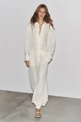 Camisa blanca a rayas verticales, de cuello redondo y manga larga con puños. Presenta cierre frontal con cordones ajustables con lazadas y abertura en la espalda ajustable con lazadas.