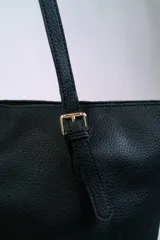 Cartera tipo shopper color negro, con textura símil cuero, doble asa de hombro con hebillas doradas y cierre superior.