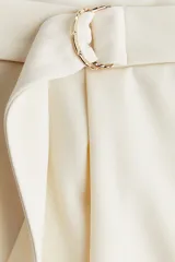 Pantalón sastrero color crudo, de tiro alto con elástico oculto en la cintura y cierre decorativo. Cinturón cosido con anillas en D de metal en un lateral. Piernas ajustadas con pinzas para estilizar la silueta.