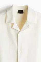Camisa de manga corta color crema, de corte holgado, con cuello resort y cierre frontal con botones.