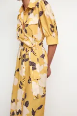 Vestido midi camisero color amarillo con estampado de flores blancas y hojas marrones. Tiene cuello solapa, mangas 3/4 con puños, cinturón de lazo ajustable y cierre frontal con botones.