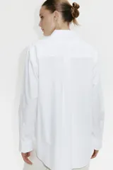 Camisa blanca de algodón con cuello clásico, abotonada adelante y corte holgado. Tiene mangas largas con puños sencillos y bajo redondeado con espalda algo más larga.