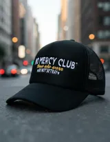 Gorra trucker negra con frente rígido y rejilla trasera. Bordado frontal con la frase "No Mercy Club" en blanco y amarillo.