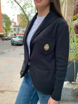 Blazer negro de tejido de punto en algodón, con solapas, dos bolsillos frontales y un escudo bordado en el pecho. Corte recto y holgado.