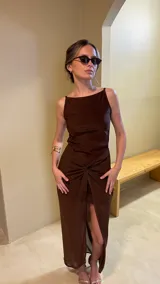 Vestido largo marrón oscuro, ajustado al cuerpo, con escote cuadrado y tirantes finos. La falda presenta un drapeado frontal anudado que crea una abertura alta.