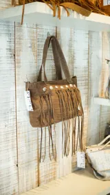 Bolso de cuero color beige con doble asa corta y correa larga extraíble. Presenta detalle de flecos largos en la parte frontal y herrajes metálicos plateados. Interior forrado con bolsillos. Marca Brava.