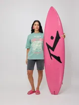 Remera blanca de algodón con estampado frontal de estilo playero con texto "Welcome to the Surf Camp" y dibujo de una casa de playa con palmeras y tablas de surf.