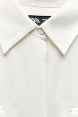 Camisa negra de satén con cuello solapa, manga larga, bolsillos con solapa en el delantero, bajo asimétrico y cierre frontal con botones.