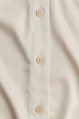 Camisa color crema de tela satinada, con escote en V, cuello clásico, cierre frontal con botones, mangas largas con puños abotonados y ruedo redondeado.