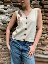 Chaleco color crema tejido en material sintético con detalle de apliques en cuero color marrón en el hombro. Tiene cuello en V, cierre frontal con botones y un bolsillo de parche en el frente.